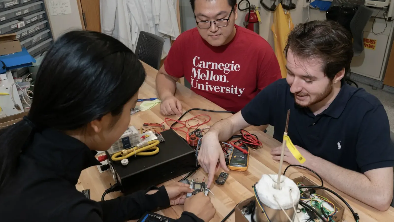 A group of three people in a workshop setting work with wires, meters and circuit components on a wooden table.